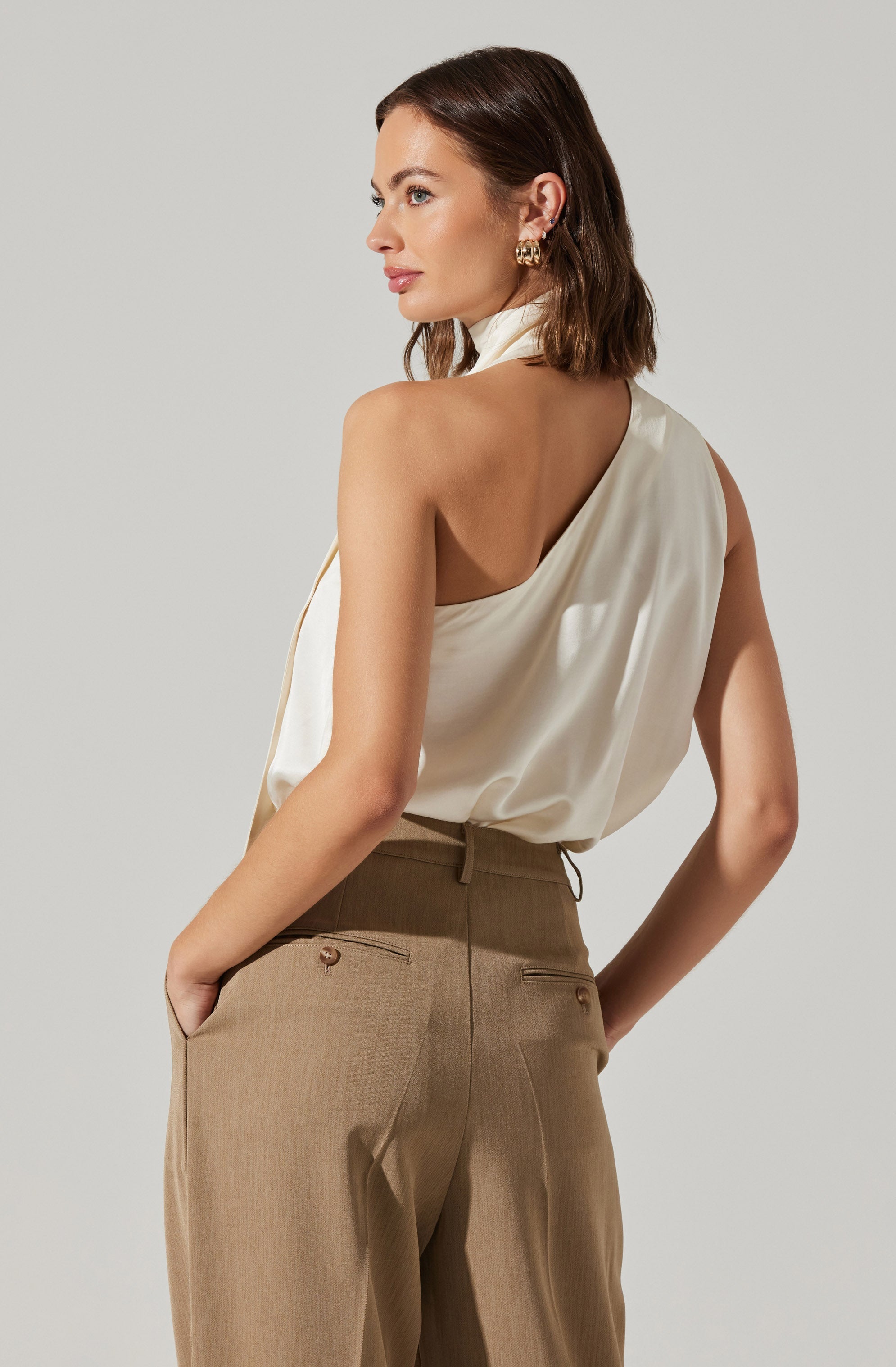 A woman is wearing a sleeveless, white satin top with a draped, asymmetrical design that exposes one shoulder and has a high collar. She has paired it with high-waisted, beige trousers that feature belt loops, side pockets, and back welt pockets.