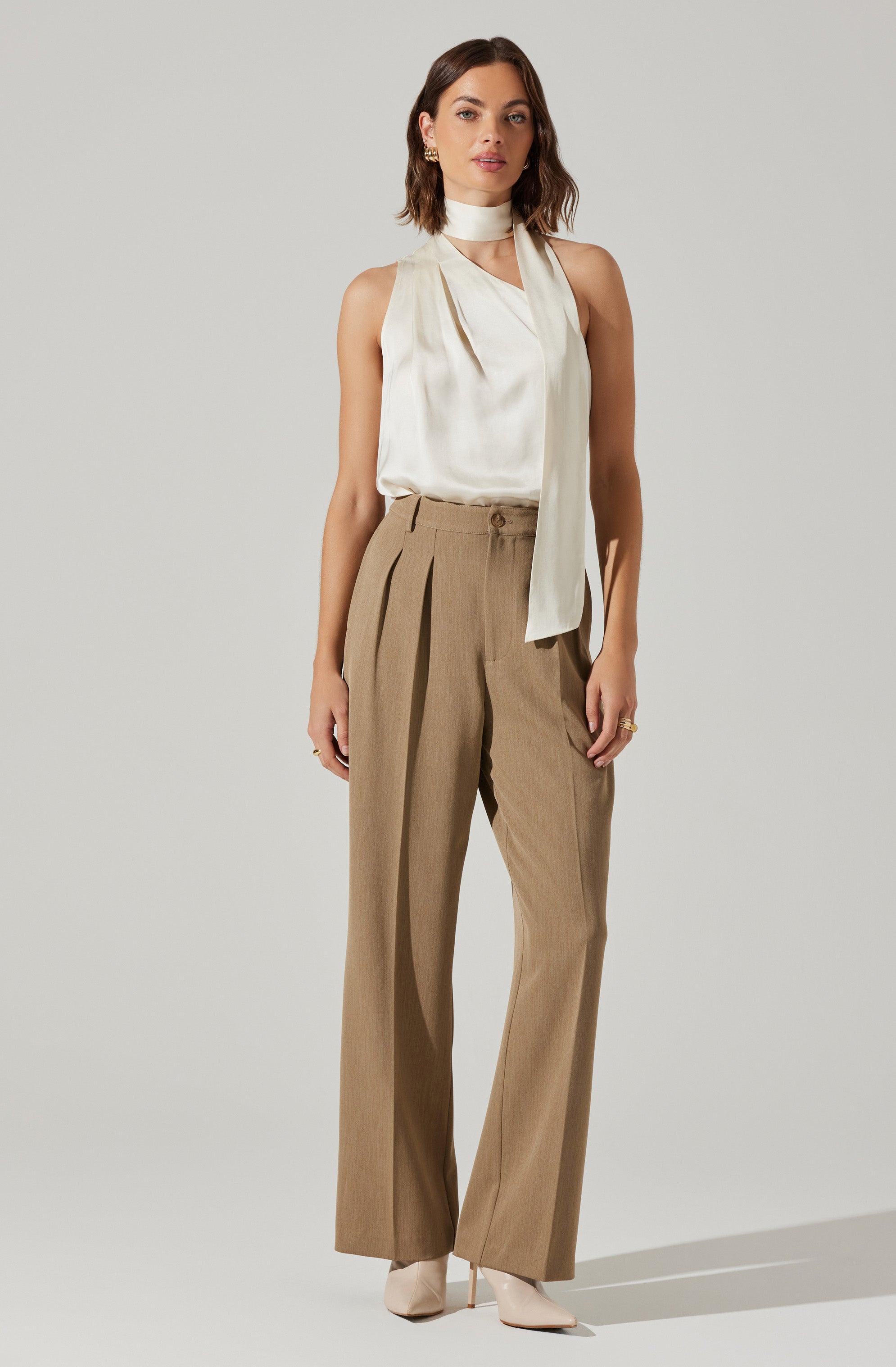 The woman is wearing a sleeveless, high-neck cream-colored blouse with a draped scarf detail around the neck. She pairs it with high-waisted, wide-leg tan trousers that feature pleats at the waist. She is also wearing ankle-high beige heels.