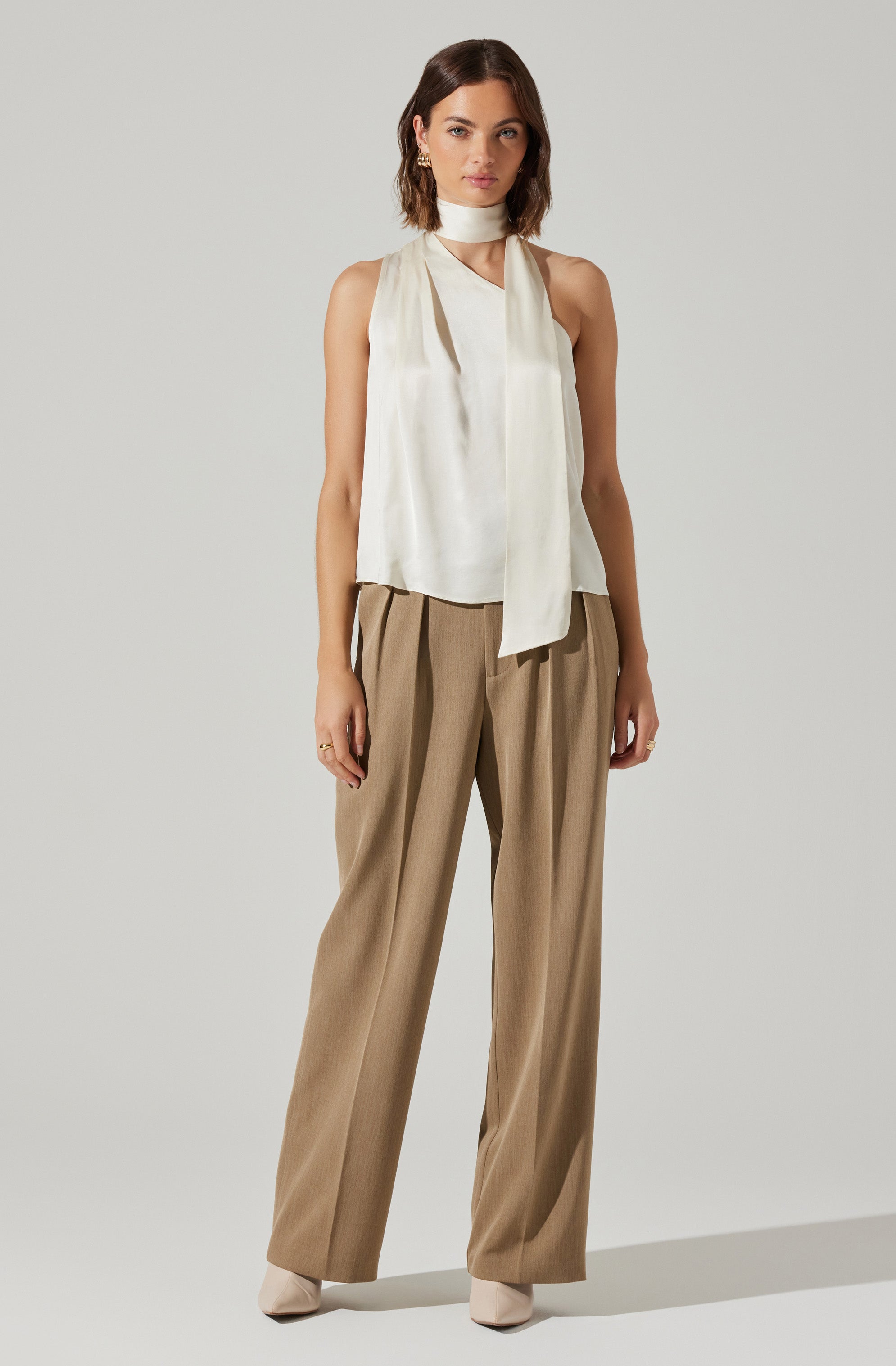 A woman is wearing a sleeveless white satin top with a high neck and a long, wide satin scarf draped around her neck. She pairs it with loose-fitting, high-waisted tan trousers that have a pleated front. She completes the look with beige pointed-toe shoes.