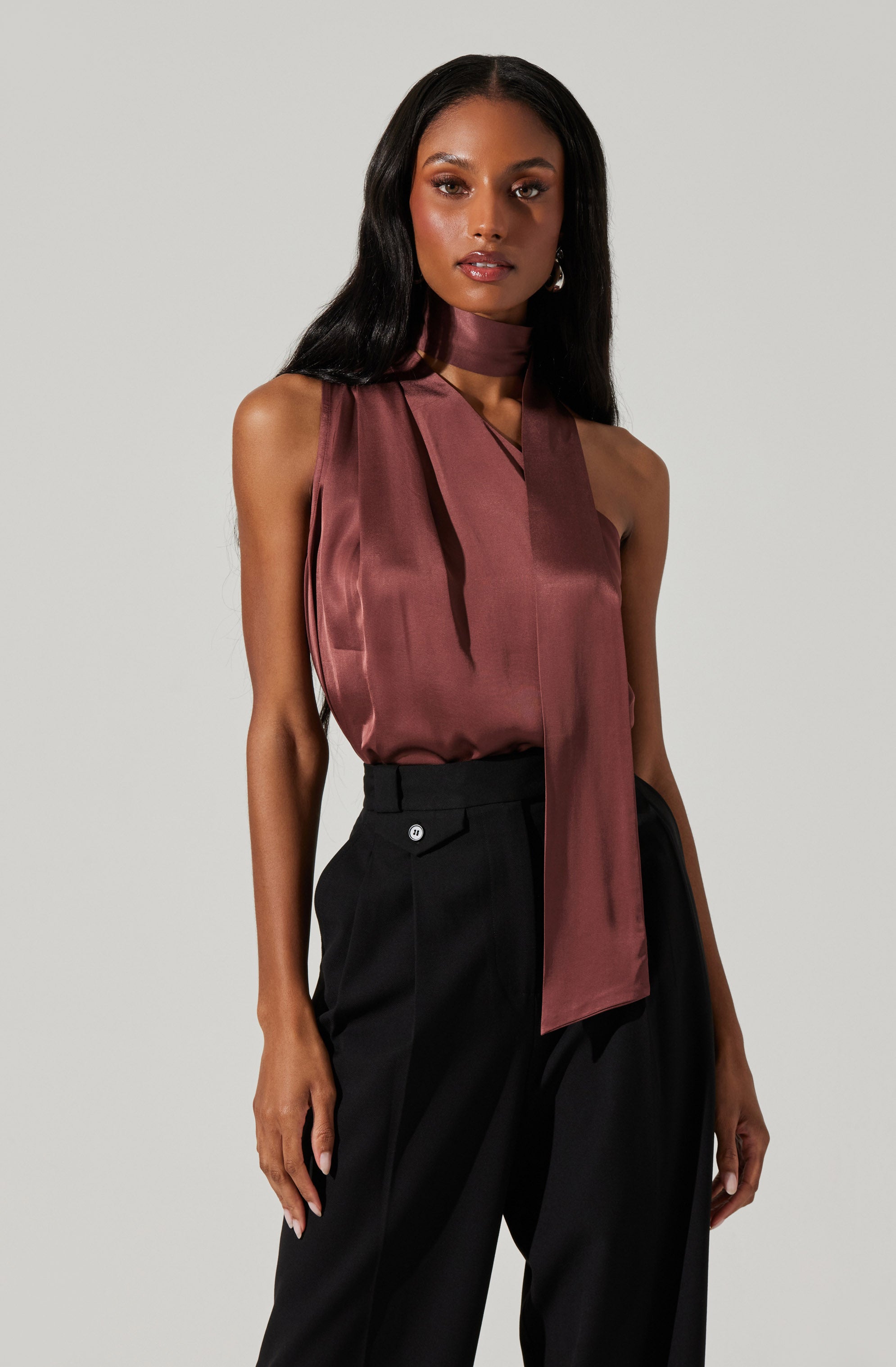 A woman is wearing a sleeveless, high-neck satin top in a deep mauve color with a long tie hanging from the neckline, paired with high-waisted black trousers.