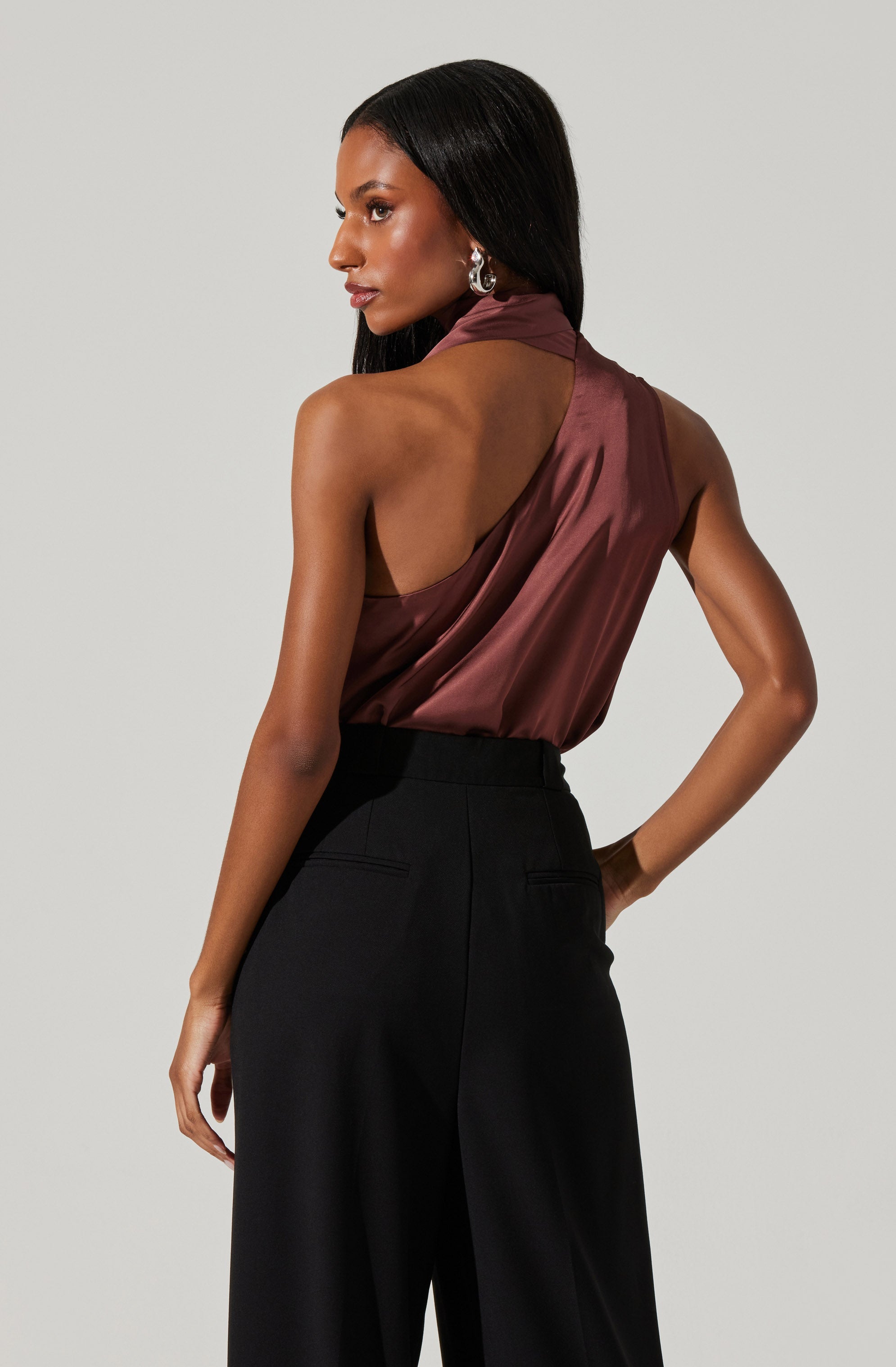 A woman is wearing a sleeveless, high-neck satin top in a deep brown or burgundy color. The top has a draped design with a halter-style neck. She pairs it with high-waisted black wide-leg trousers.