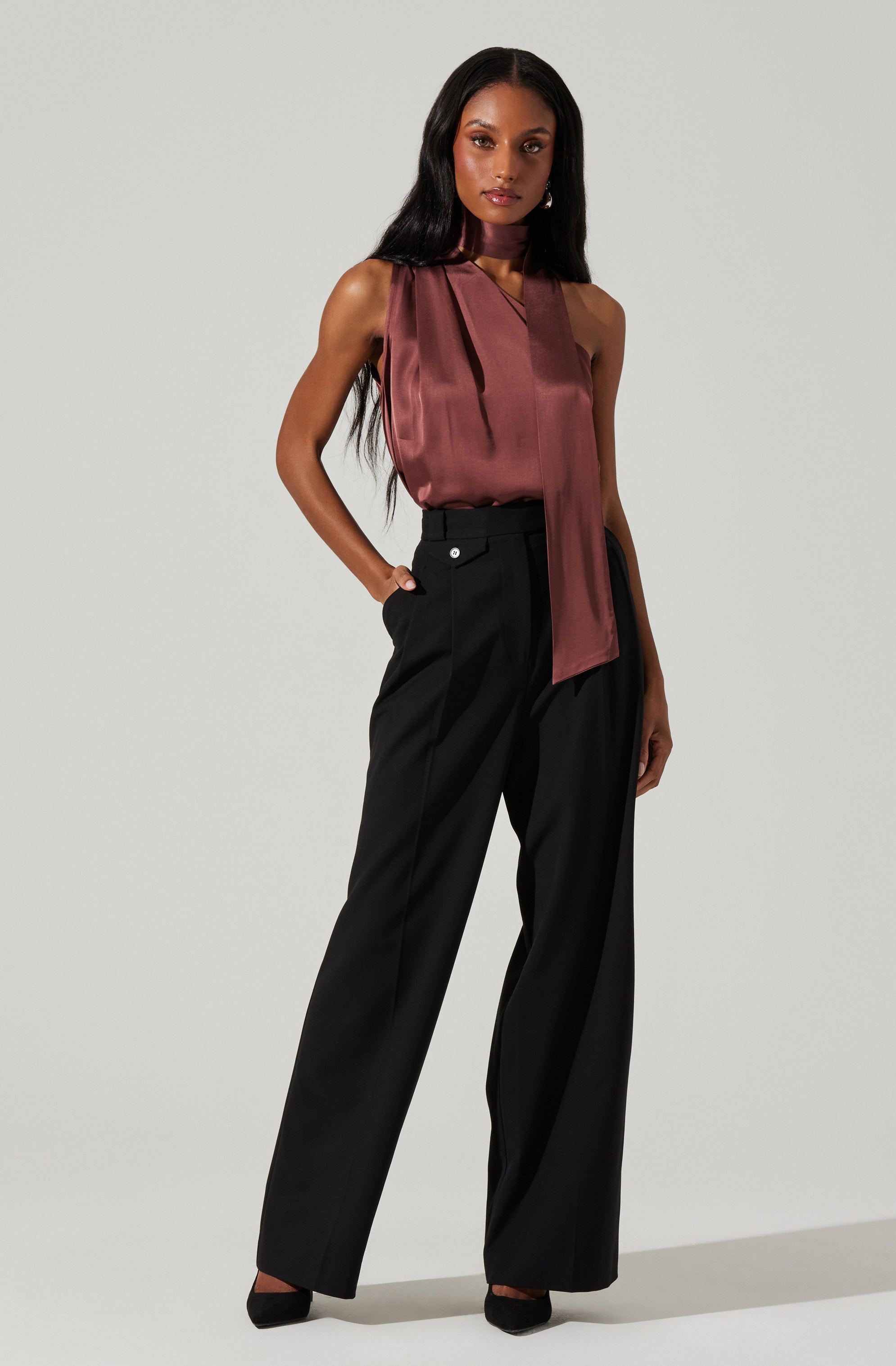 A woman is wearing a sleeveless satin top in a deep mauve or burgundy color with a large, draped tie detail hanging down from the neckline. She pairs the top with high-waisted, wide-leg black trousers that have a tailored fit. She completes the look with black pointed-toe shoes and small, subtle earrings.