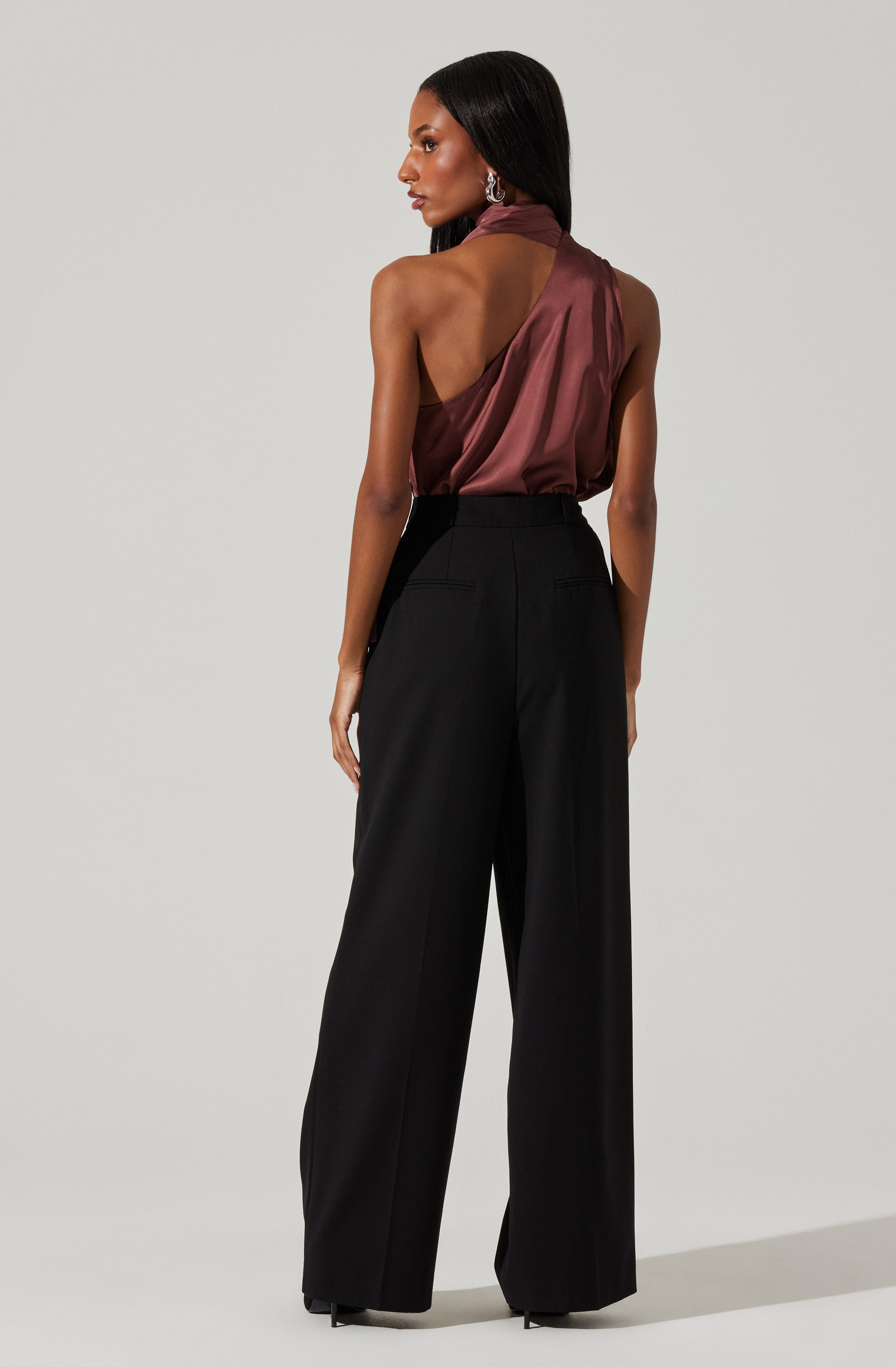 A woman is wearing a sleeveless, high-neck top made of shiny, satin-like fabric in a rich, deep mauve color. The top has a draped, asymmetrical design that reveals her shoulders and part of her back. She pairs it with high-waisted, wide-leg black trousers with a tailored fit.