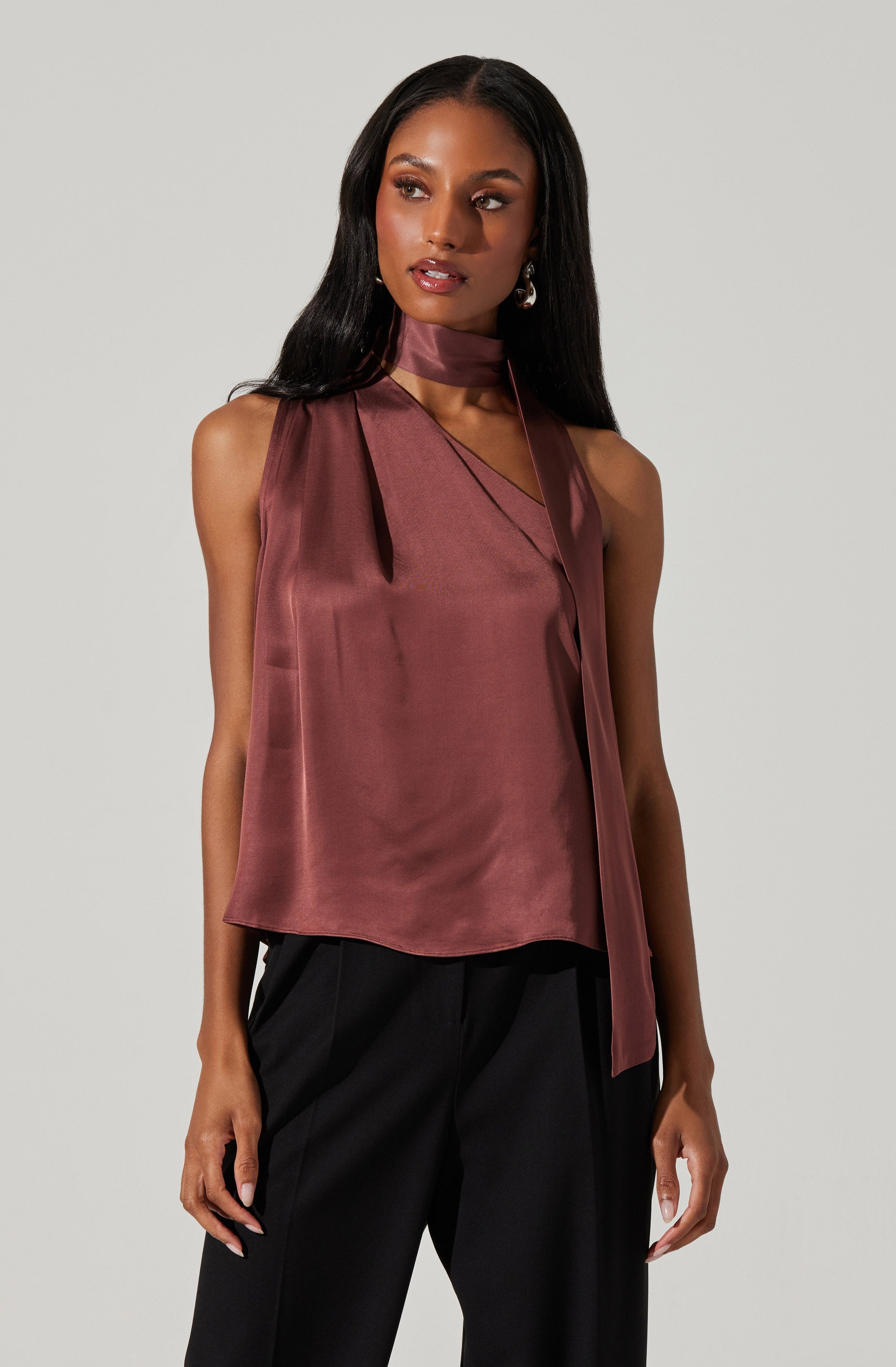 A woman is wearing a sleeveless, satin-like top in a dusty mauve color. The top features a high neck with a matching fabric tie that drapes down the front. She pairs it with black trousers.