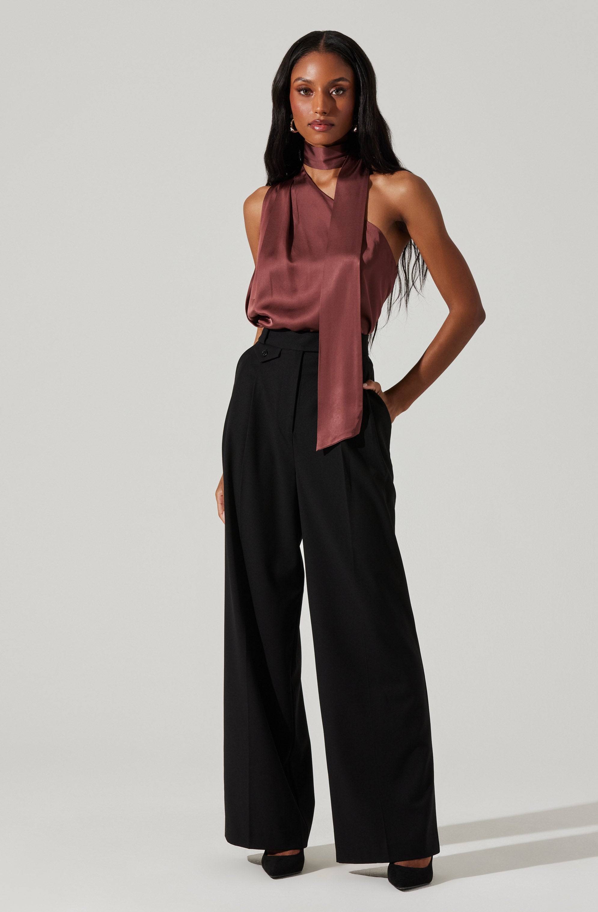 A woman is wearing a sleeveless, satin-like, burgundy top with a matching long necktie. She pairs it with high-waisted, wide-leg black trousers and black shoes.