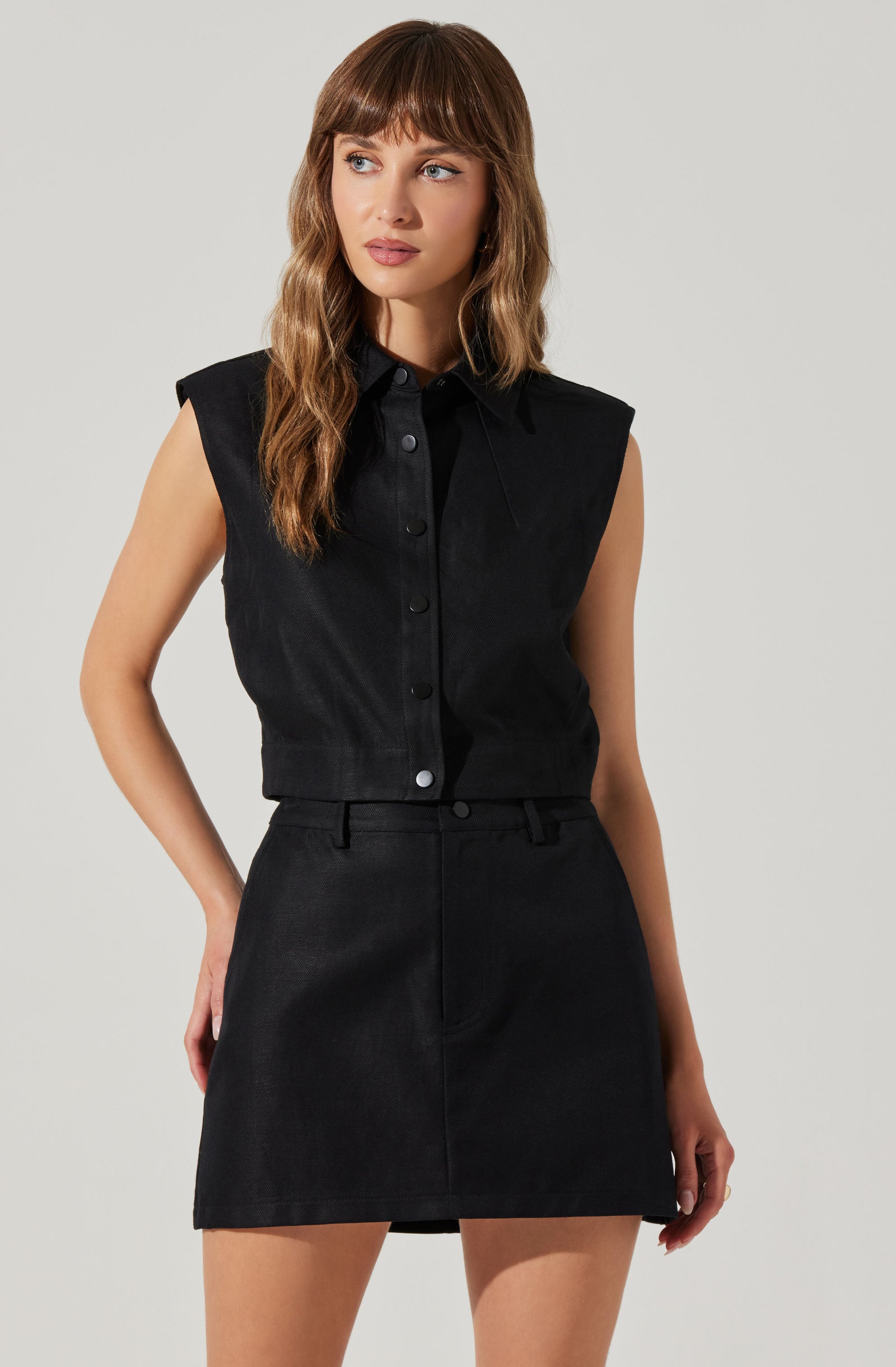 A woman is wearing a sleeveless black button-up shirt with a collar, paired with a black high-waisted skirt. The shirt has shoulder pads, giving it a structured appearance.
