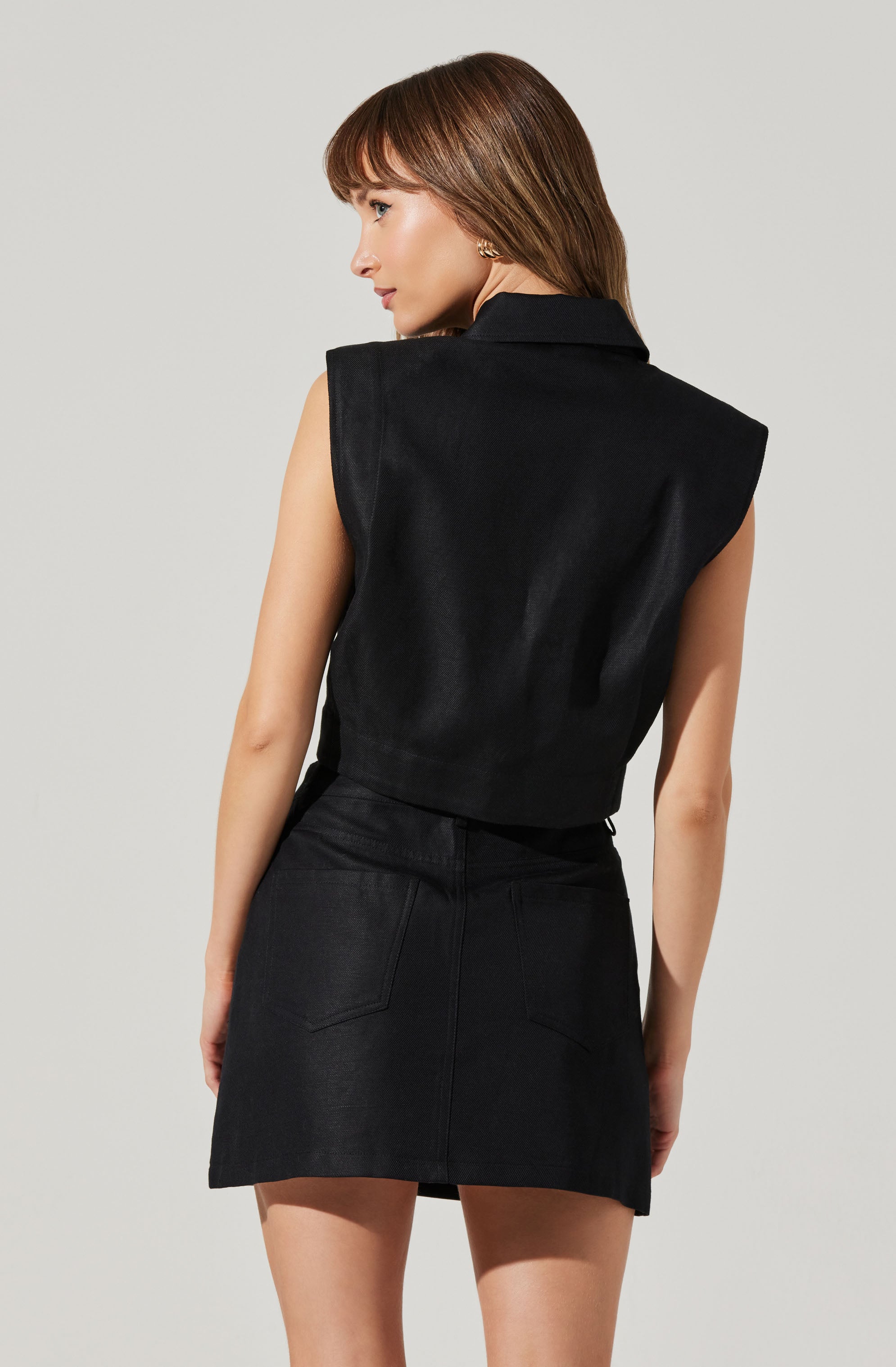 A woman is wearing a sleeveless, black collared vest with a structured shoulder design and a slightly loose fit. She pairs it with a black denim skirt that has a high waist and two back pockets.