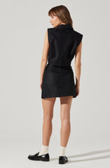 A woman is wearing a sleeveless black top with padded shoulders and a matching black mini skirt. She is also wearing white socks and shiny black loafers.