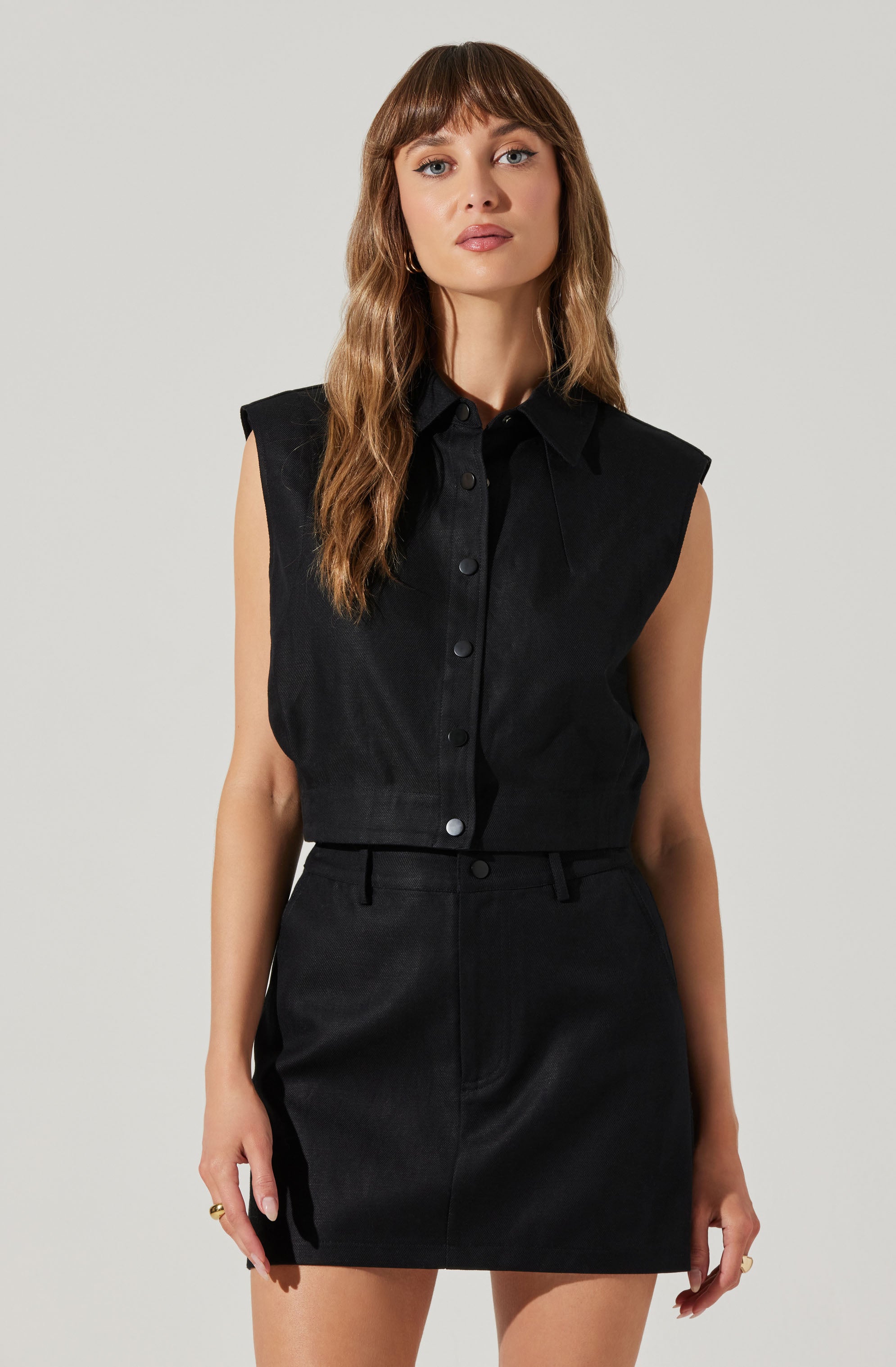 A woman is wearing a sleeveless black button-up shirt with broad shoulder pads, paired with a black high-waisted skirt that has belt loops. The outfit is monochromatic and has a sleek, minimalist style.