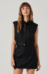 A woman is wearing a sleeveless black button-up shirt with broad shoulder pads, paired with a black high-waisted skirt that has belt loops. The outfit is monochromatic and has a sleek, minimalist style.