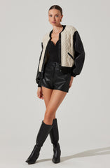 A woman is wearing a black V-neck top with a fitted silhouette, black high-waisted shorts made of faux leather, and knee-high black leather boots with a slight heel. She has a cream and black varsity-style jacket with a cable-knit pattern on the body, black leather sleeves, and black trim along the collar, cuffs, and hem.
