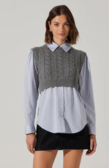 A woman is wearing a light blue, long-sleeved, button-up shirt with thin vertical white and blue stripes. Over the shirt, she is wearing a gray, sleeveless, knitted sweater vest with cable knit patterns. She is also wearing a black skirt.