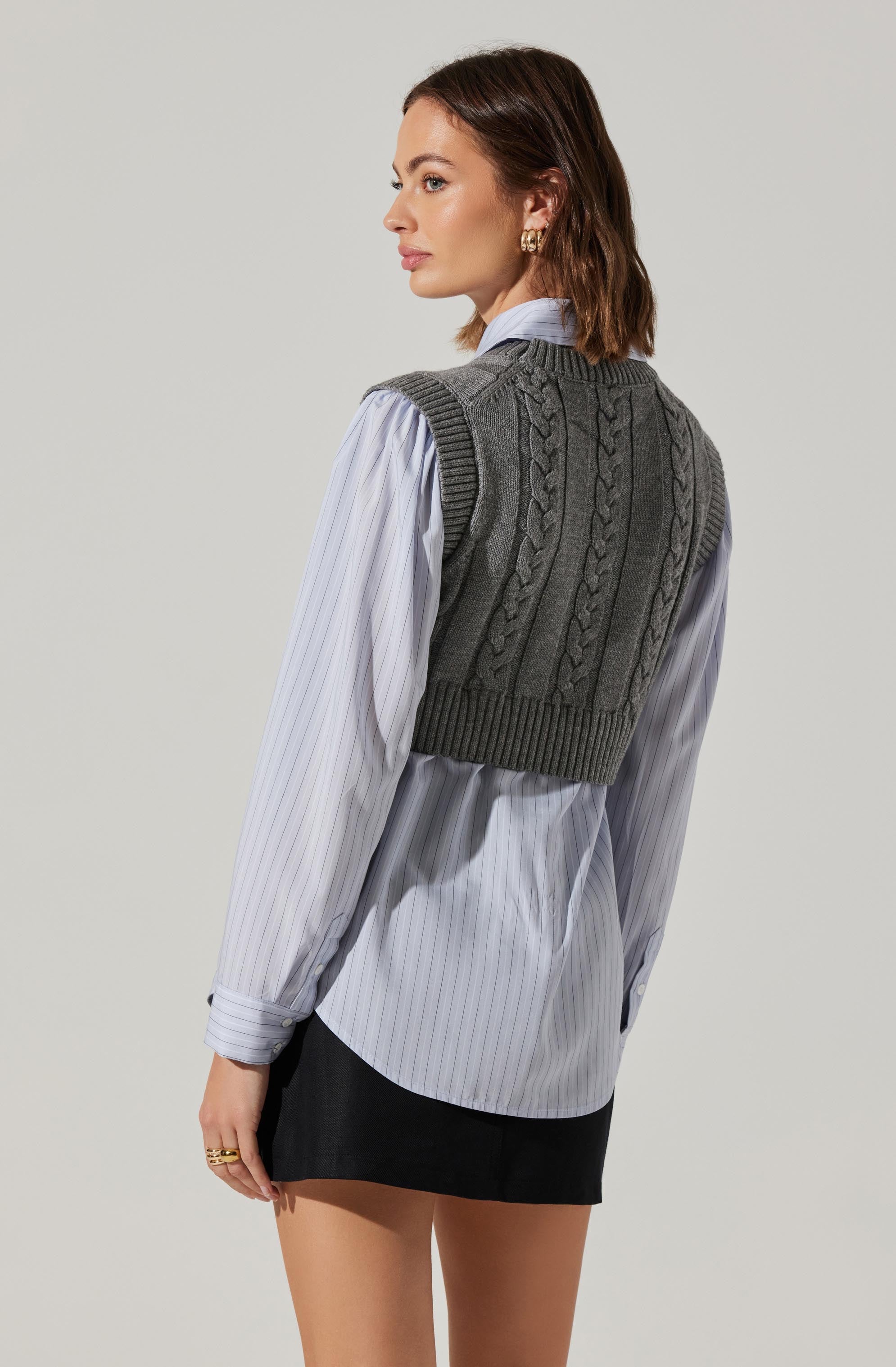A woman is wearing a sleeveless, dark gray cable-knit sweater vest layered over a long-sleeve, light blue and white vertically striped button-up shirt.