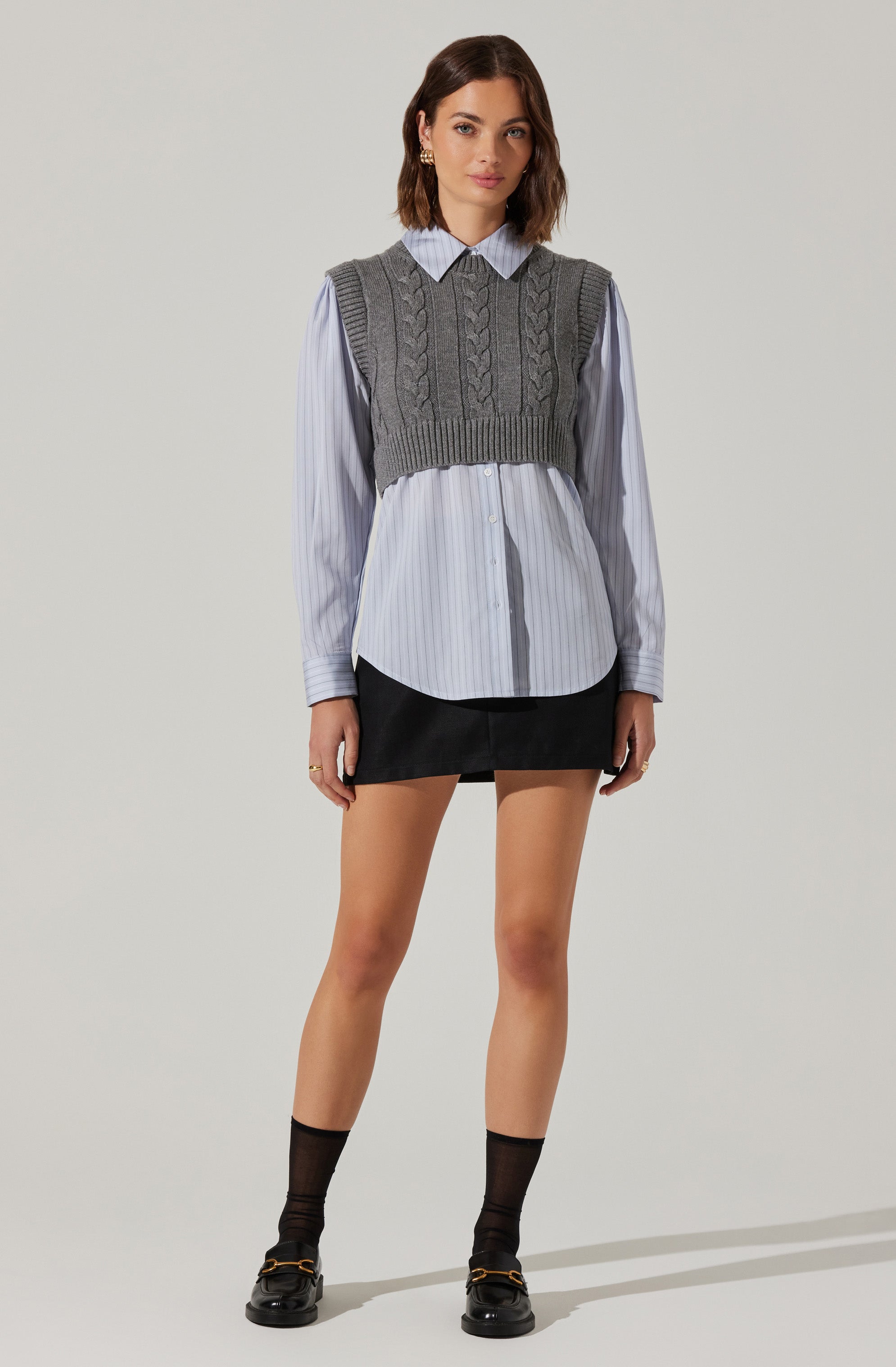 A woman is wearing a light blue, long-sleeve, vertically striped shirt with a collar and button-down front. Over the shirt, she has on a gray, sleeveless, cable-knit cropped sweater vest. She is also wearing a black mini skirt, black ankle socks, and black loafers with gold hardware.