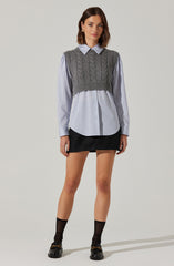 A woman is wearing a light blue, long-sleeve, vertically striped shirt with a collar and button-down front. Over the shirt, she has on a gray, sleeveless, cable-knit cropped sweater vest. She is also wearing a black mini skirt, black ankle socks, and black loafers with gold hardware.