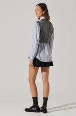 A woman is wearing a light blue, long-sleeved, button-down shirt with vertical white stripes. Over the shirt, she has on a dark gray, ribbed, sleeveless knitted vest with cable knit pattern. She is also wearing a short black skirt, black ankle socks, and black loafers with gold buckle accents.