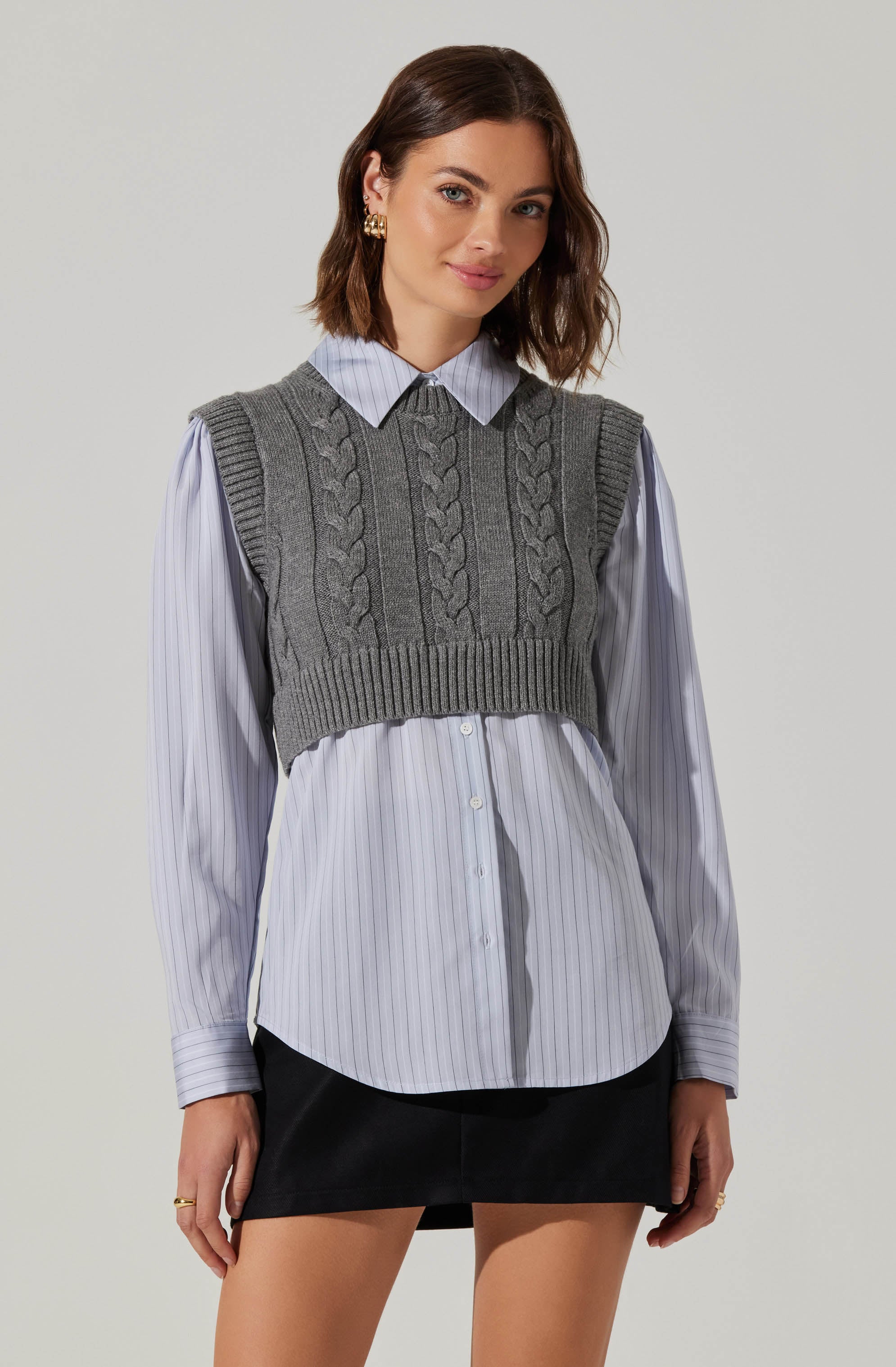 A young woman is wearing a light blue collared shirt with thin vertical white stripes, buttoned down the front. Over the shirt, she has on a gray knit sleeveless sweater vest featuring braided cable patterns on the front and ribbed details at the hem and armholes. She pairs the top with a black skirt.