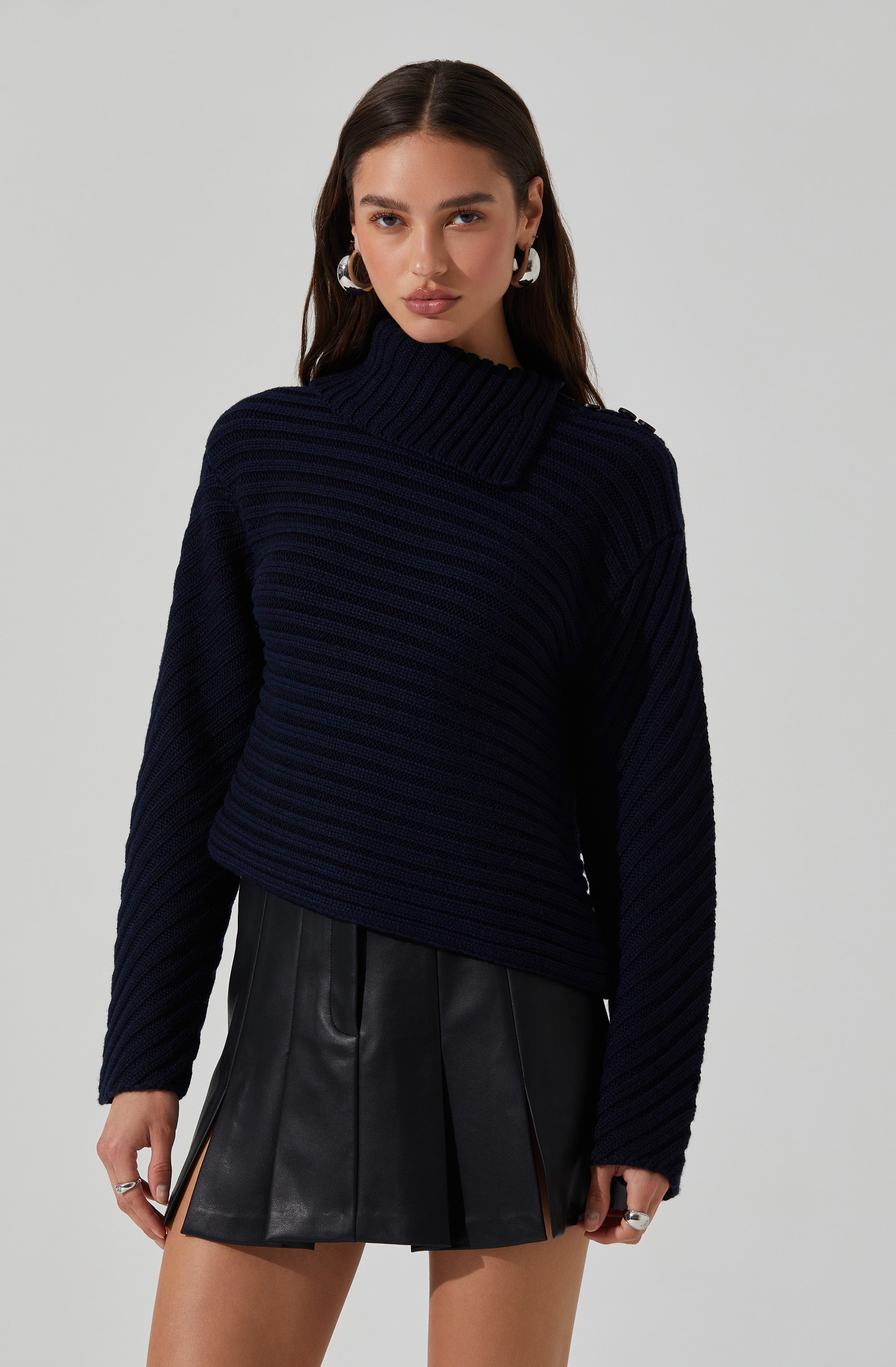 A young woman is wearing a dark navy blue, chunky knit sweater with a wide, ribbed turtleneck that extends across one shoulder. She pairs it with a black, high-waisted leather mini skirt featuring front pleats and side zipper details. She accessorizes with silver hoop earrings and rings.
