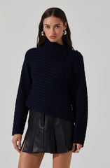 A young woman is wearing a dark navy blue, chunky knit sweater with a wide, ribbed turtleneck that extends across one shoulder. She pairs it with a black, high-waisted leather mini skirt featuring front pleats and side zipper details. She accessorizes with silver hoop earrings and rings.