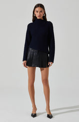A woman is wearing a dark navy, ribbed, turtleneck sweater with asymmetric shoulder details, paired with a black leather mini skirt featuring pleats. She completes her outfit with black pointed-toe heels with embellishments.