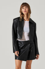 A woman is wearing a black leather cropped jacket with a pointed collar, long sleeves, and a front zipper. She has on a white T-shirt underneath and is also wearing matching black leather shorts with a visible button and zipper closure.