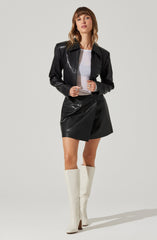 A woman is wearing a shiny black leather jacket with a collar, paired with a white t-shirt underneath. She has on a matching black leather skirt with an asymmetrical wrap design. She is also wearing tall, white knee-high boots with a slight heel.