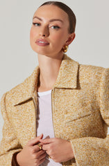 A woman is wearing a textured, multicolored tweed jacket in shades of yellow, orange, and cream. The jacket has a collar and a front zipper. Underneath, she has on a white T-shirt. She is accessorized with large, gold hoop earrings and gold rings on her fingers.