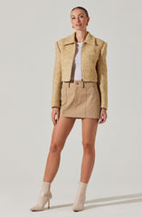 A woman is wearing a cropped, textured, beige jacket with a collar and long sleeves. She has on a white top underneath, a beige mini skirt with front seams and belt loops, and beige high-heeled ankle boots with pointed toes.