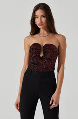 A woman is wearing a strapless, fitted top covered in dark red sequins with a central cut-out detail. She is also wearing high-waisted black trousers. She accessorizes with a chunky silver chain necklace, earrings, and rings on her fingers.