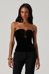 A woman is wearing a strapless black velvet top with a central cutout and ruched detailing. She pairs it with black pants and accessorizes with gold jewelry, including a chunky bracelet, rings, and a necklace.