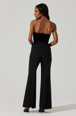 A woman is wearing a strapless black top made of a textured fabric, paired with high-waisted black wide-leg pants. She is also wearing black high heels, a gold chain necklace, and hoop earrings. Her hair is long and straight.