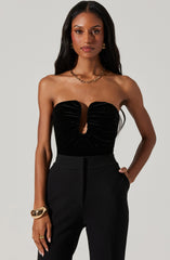 A woman is wearing a strapless black top with a keyhole cutout in the center, featuring gathered fabric detail that radiates outward from the cutout. She pairs the top with high-waisted black trousers. She accessorizes with a chunky gold chain necklace, gold hoop earrings, a gold bracelet, and rings.