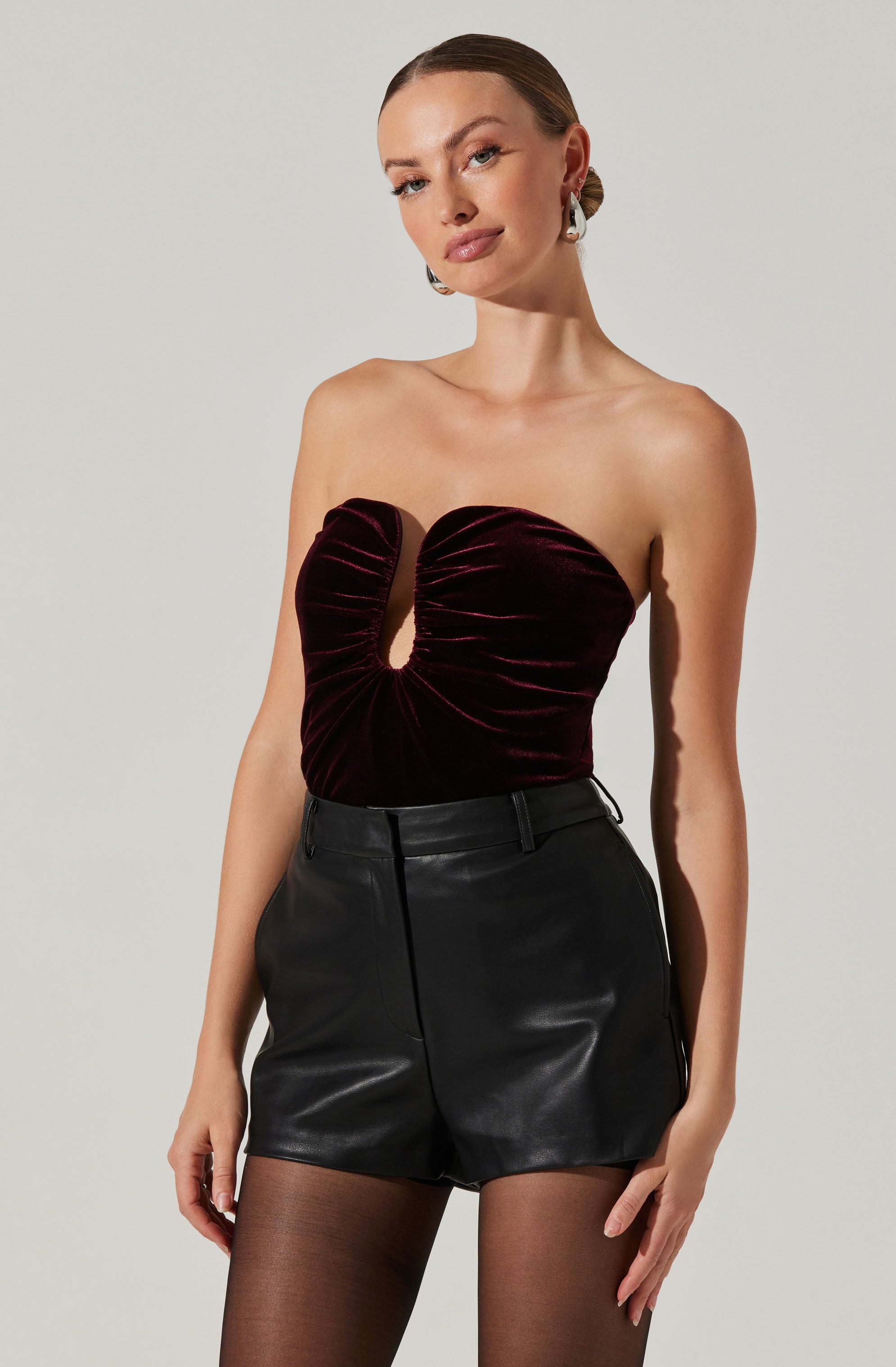 A woman is wearing a strapless top made of dark burgundy velvet fabric with a keyhole cutout at the center. She pairs it with high-waisted black leather shorts and sheer black tights.