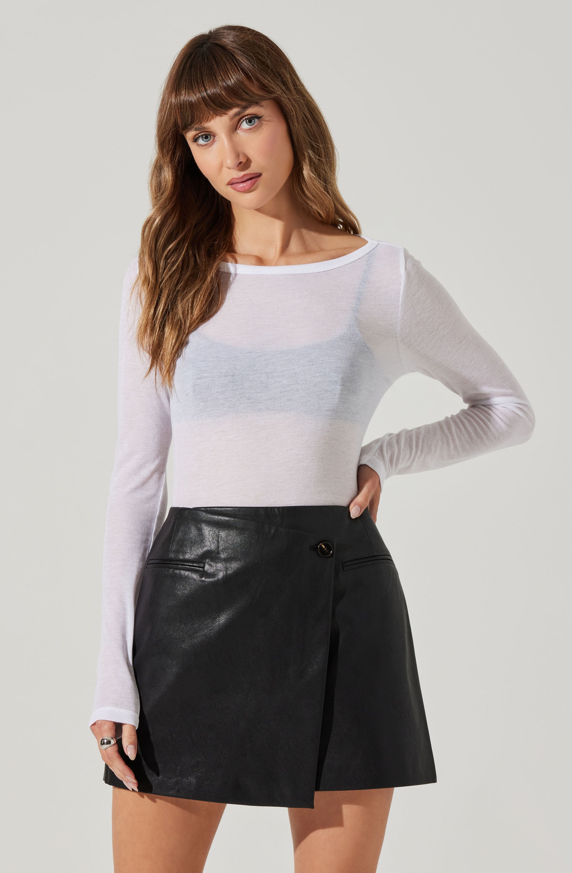 A woman is wearing a long-sleeve, white, semi-sheer top that is fitted and slightly see-through. She has paired it with a high-waisted, black mini skirt made of faux leather, featuring a wrap-style design with a side button closure and two small front pockets.