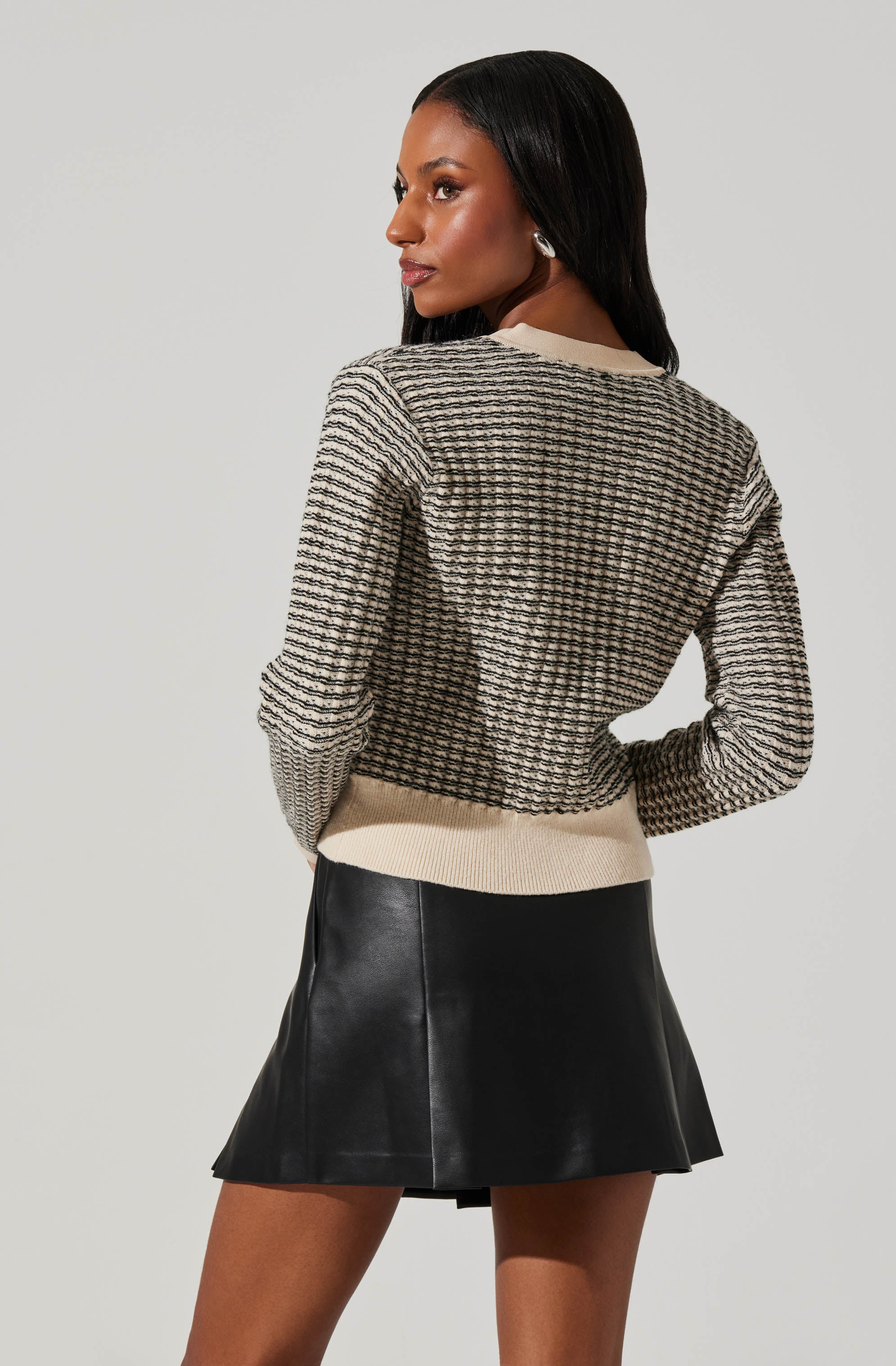 A woman is wearing a long-sleeved, knit sweater with a textured, checkered pattern in neutral tones. The sweater has a beige ribbed hem and collar. She is also wearing a black, leather-like A-line mini skirt.