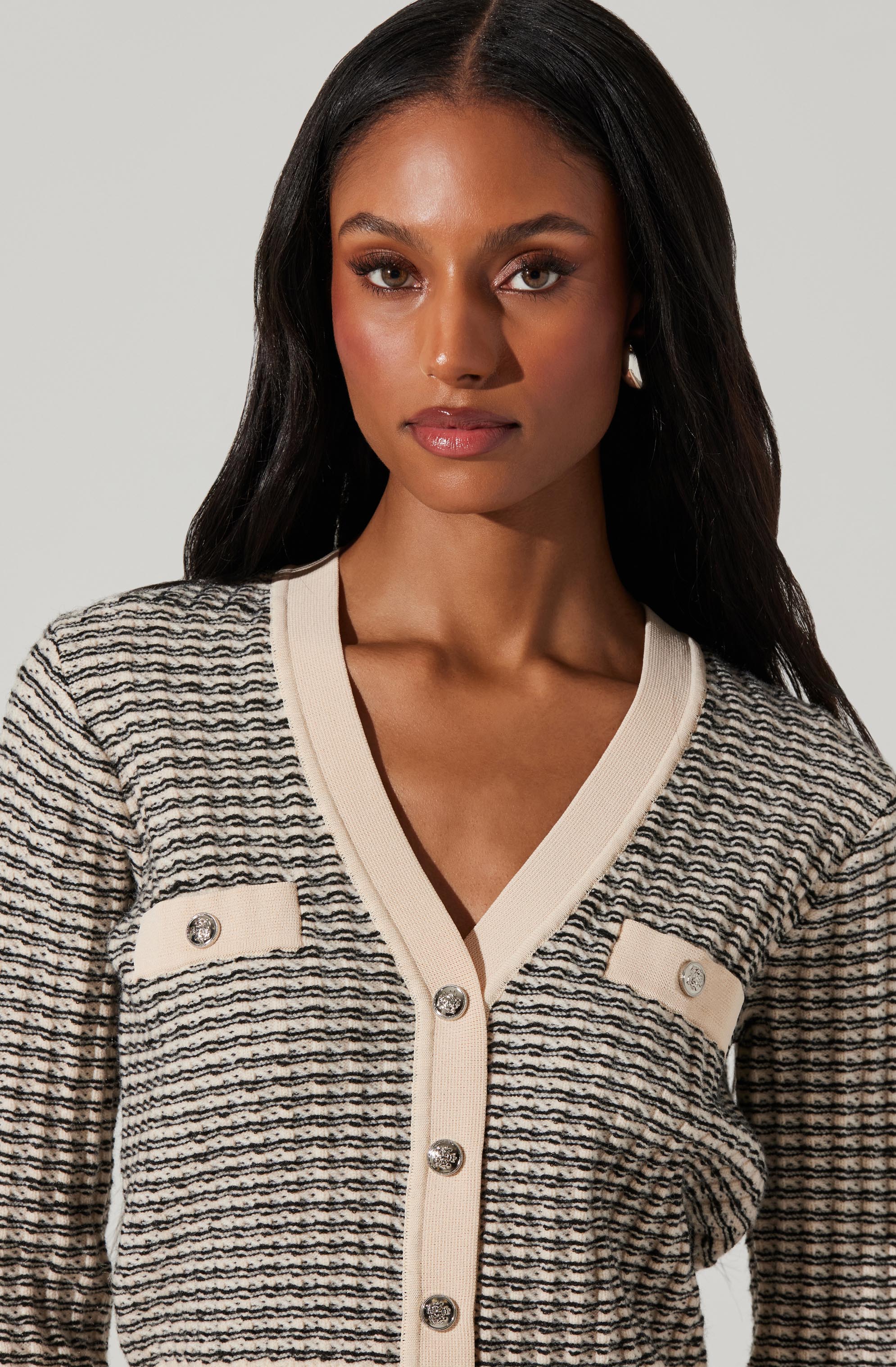 A woman is wearing a cream-colored cardigan with horizontal black and gray stripes. The cardigan features a V-neckline, cream-colored trim, and decorative buttons with diamond-like embellishments on the front pockets and center.