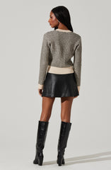A woman is wearing a long-sleeved knit sweater with a black and white checkered pattern and ribbed beige cuffs and waistband. She pairs it with a black leather mini skirt and tall black leather boots with block heels.