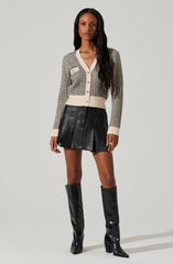 A woman is wearing a beige and black patterned cardigan with a v-neckline, long sleeves, and buttons down the front. She pairs it with a black leather pleated mini skirt and knee-high black leather boots with pointed toes and block heels.