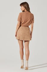 A woman is wearing a loose-fitting, short-sleeved, camel-colored top with a high collar. She pairs it with a textured, peach-colored skirt that has a slightly flared hem. She completes her outfit with a pair of light-colored, high-heeled ankle boots.