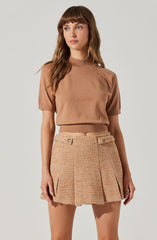 A woman is wearing a brown short-sleeve top with a high neckline and a ribbed waistband. She pairs it with a beige textured mini skirt featuring decorative buttons at the waistband and pleats along the front.