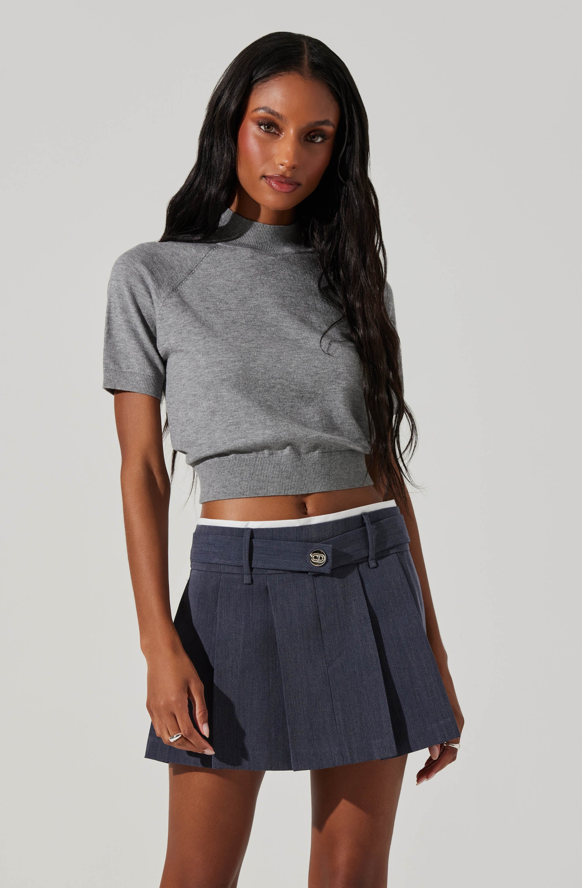 A woman is wearing a gray short-sleeve cropped sweatshirt with a high neckline and elastic waistband, paired with a navy pleated skirt featuring a front button and belt loops.