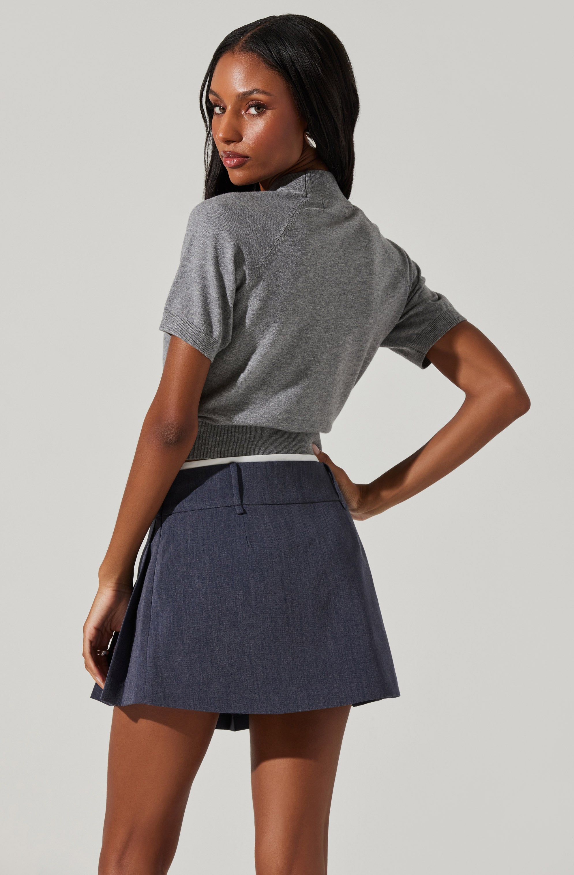 A woman is wearing a short-sleeved, heather gray t-shirt tucked into a dark blue pleated skirt with a subtle texture, and a dark waistband.