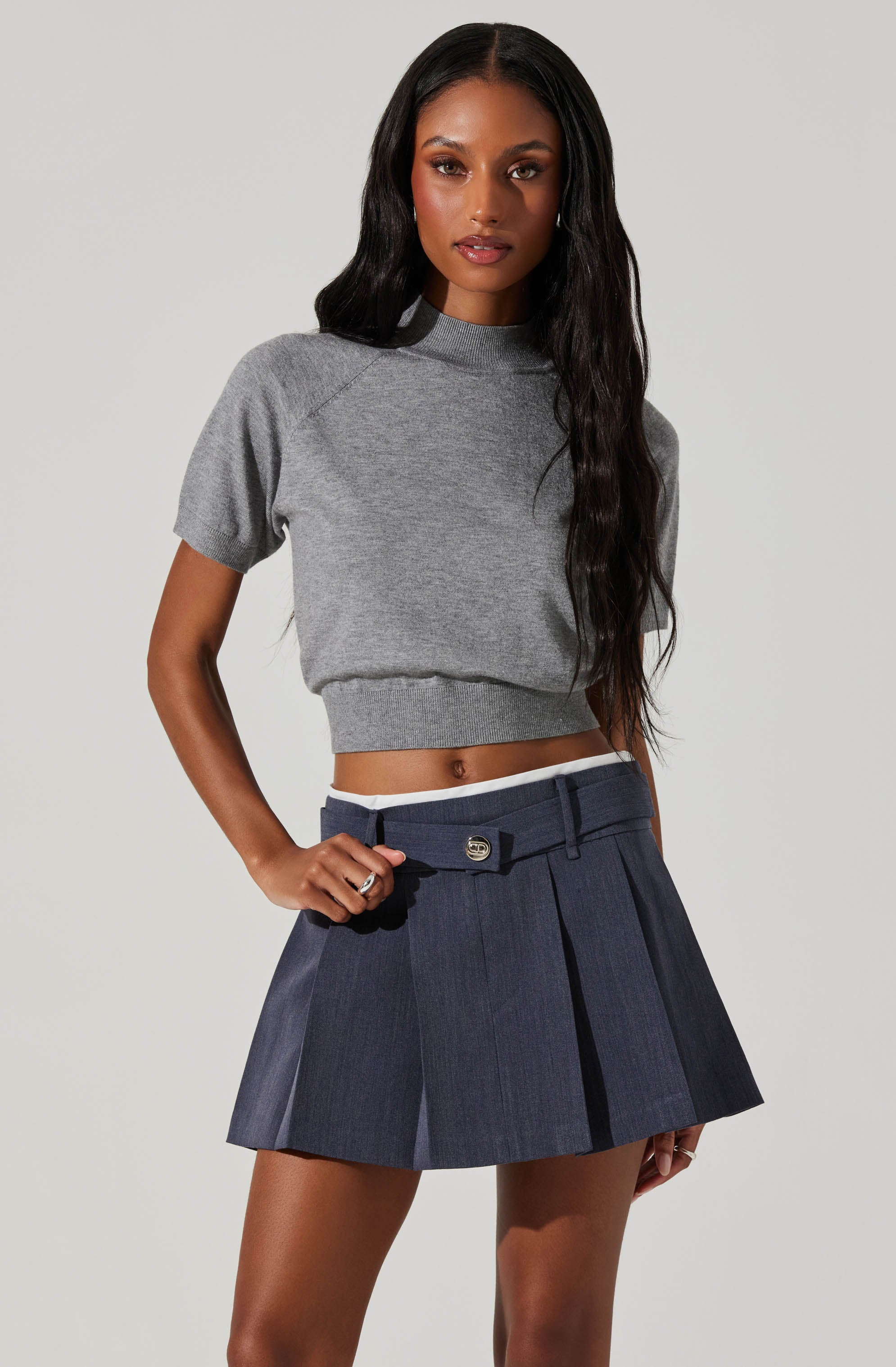 A woman is wearing a gray, short-sleeved, cropped sweatshirt with a high neckline and a wide ribbed hem. She is also dressed in a navy blue pleated skirt with a belt loop, front pleats, and a small decorative button at the waistband.