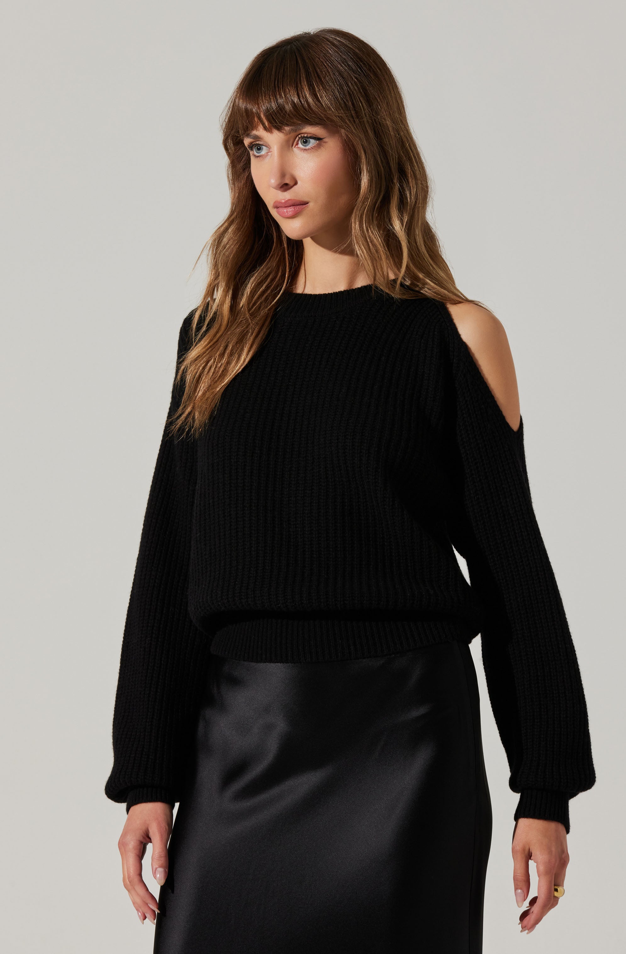 A woman is wearing a black, knitted sweater with shoulder cut-outs that reveal her skin. The sweater has a textured pattern and long sleeves with ribbed cuffs. She is also dressed in a black, shiny, satin or leather-like skirt that reaches to her knees.