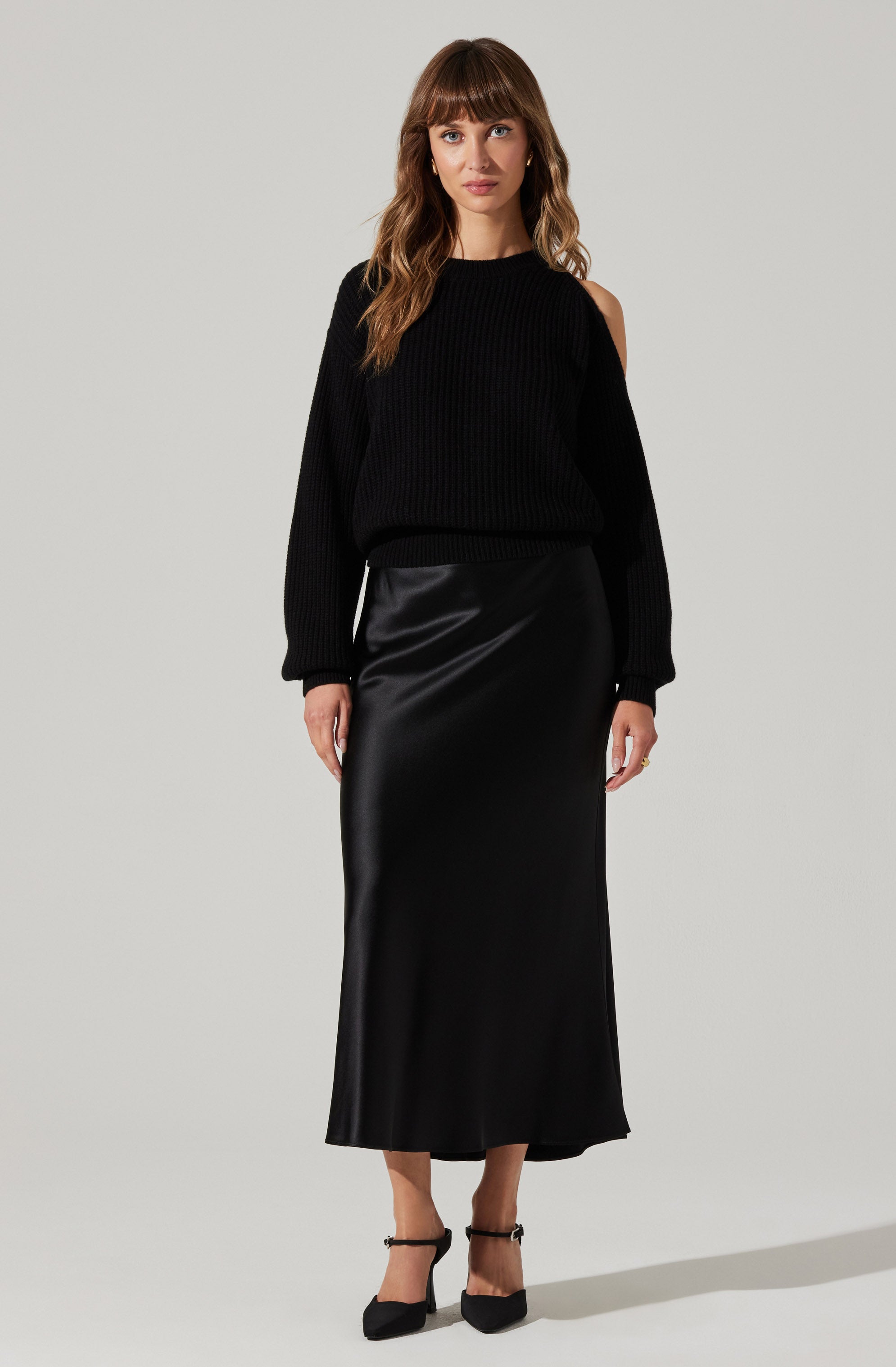A woman is wearing a black ribbed knit sweater with one shoulder exposed and long sleeves, paired with a long, silky black skirt that reaches her ankles. She is also wearing black high-heeled shoes with ankle straps.