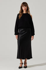 A woman is wearing a black ribbed knit sweater with one shoulder exposed and long sleeves, paired with a long, silky black skirt that reaches her ankles. She is also wearing black high-heeled shoes with ankle straps.