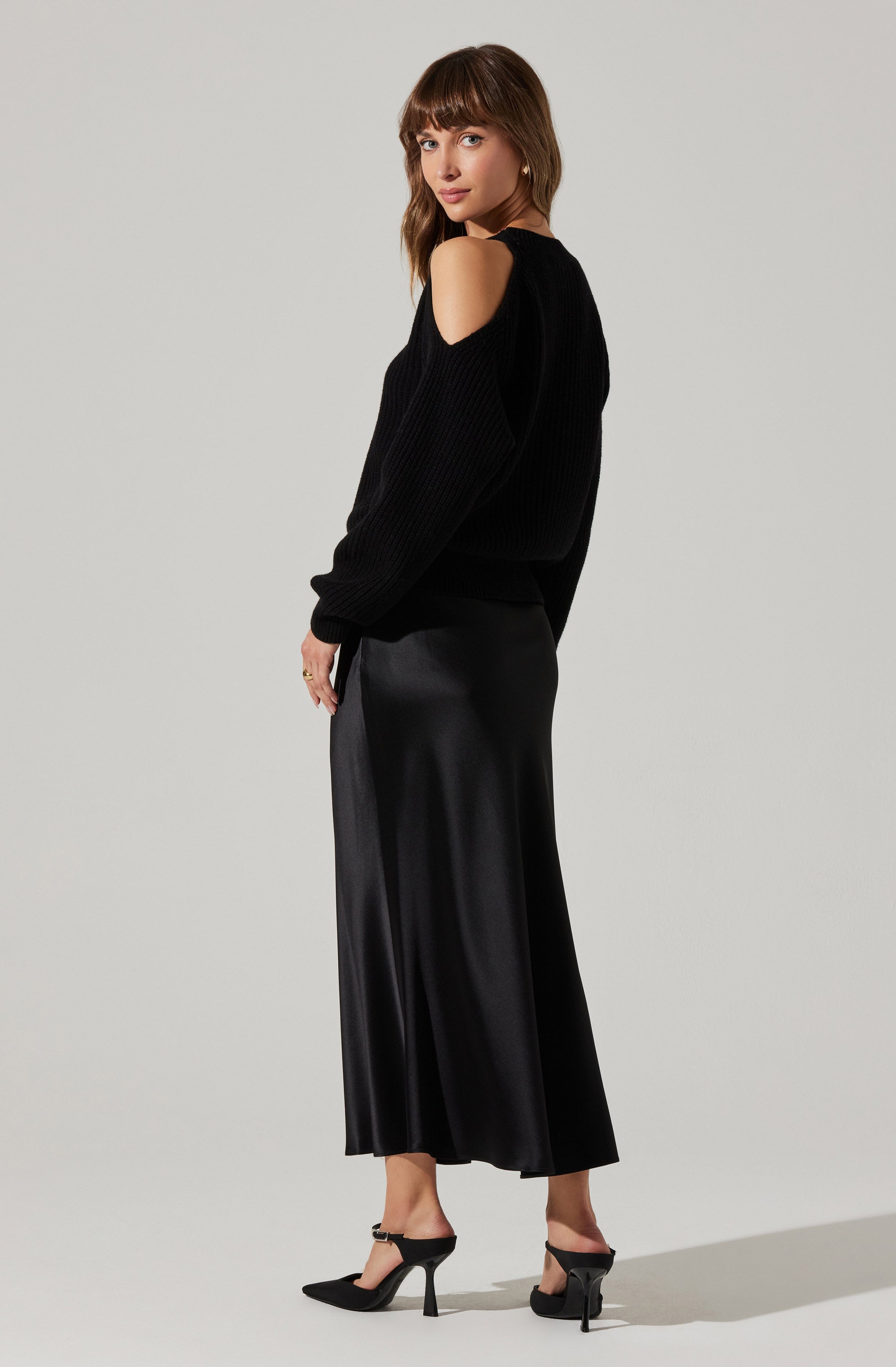 A woman is wearing a black sweater with cut-out shoulder details, featuring long sleeves and a ribbed texture. She pairs it with a long, flowy, black satin skirt that reaches her ankles and has a slight sheen. She completes her outfit with black high-heeled shoes with ankle straps.