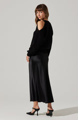 A woman is wearing a black sweater with cut-out shoulder details, featuring long sleeves and a ribbed texture. She pairs it with a long, flowy, black satin skirt that reaches her ankles and has a slight sheen. She completes her outfit with black high-heeled shoes with ankle straps.