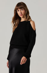 A woman is wearing a black, chunky-knit sweater with shoulder cut-outs revealing her shoulders. She pairs the sweater with a black, shiny, satin-like skirt.