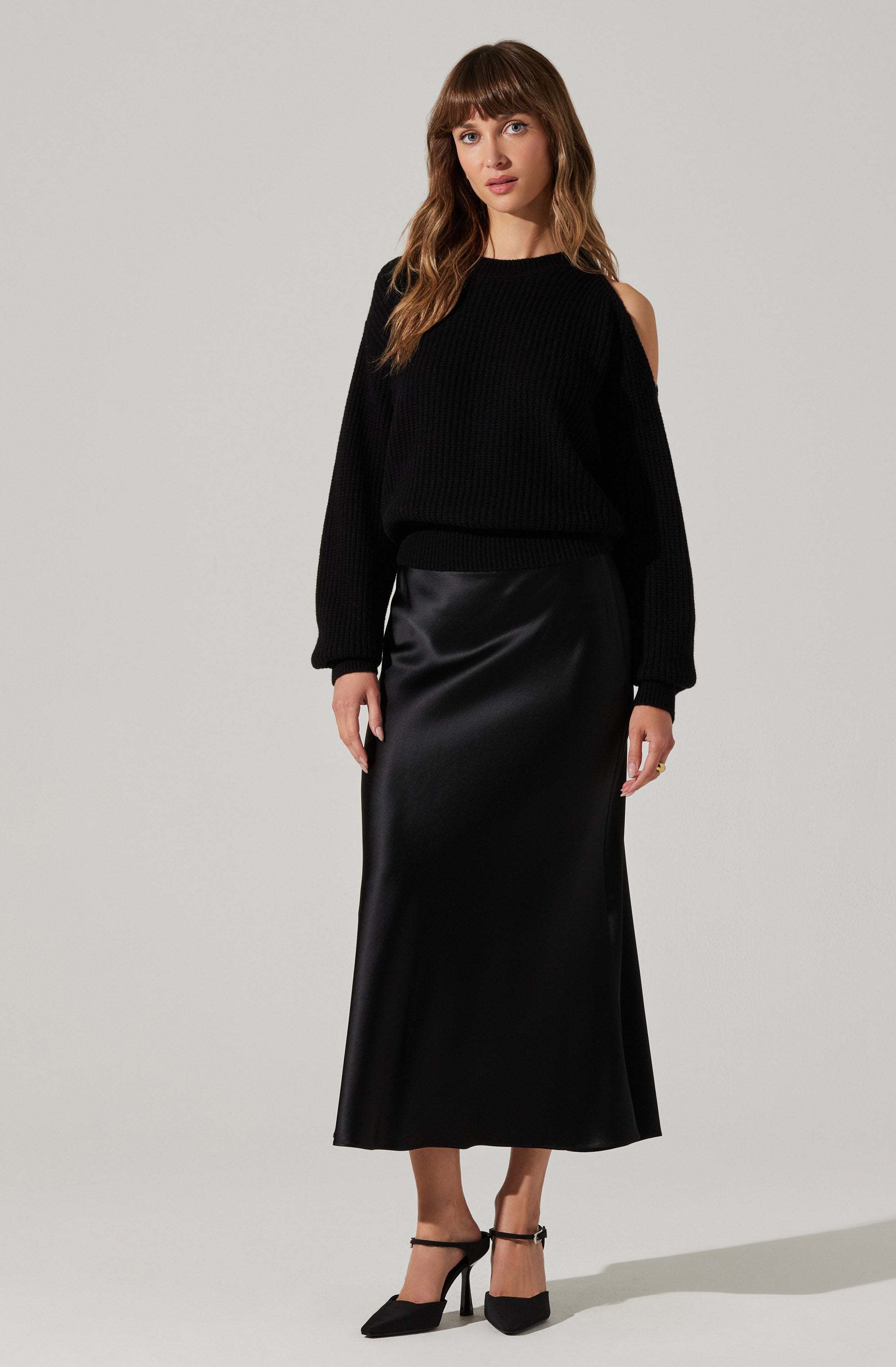 A woman is wearing a black, long-sleeved, ribbed sweater with a cold shoulder detail on the left side. She pairs it with a high-waisted, shiny satin, ankle-length black skirt. She completes her outfit with black high-heeled shoes featuring ankle straps.